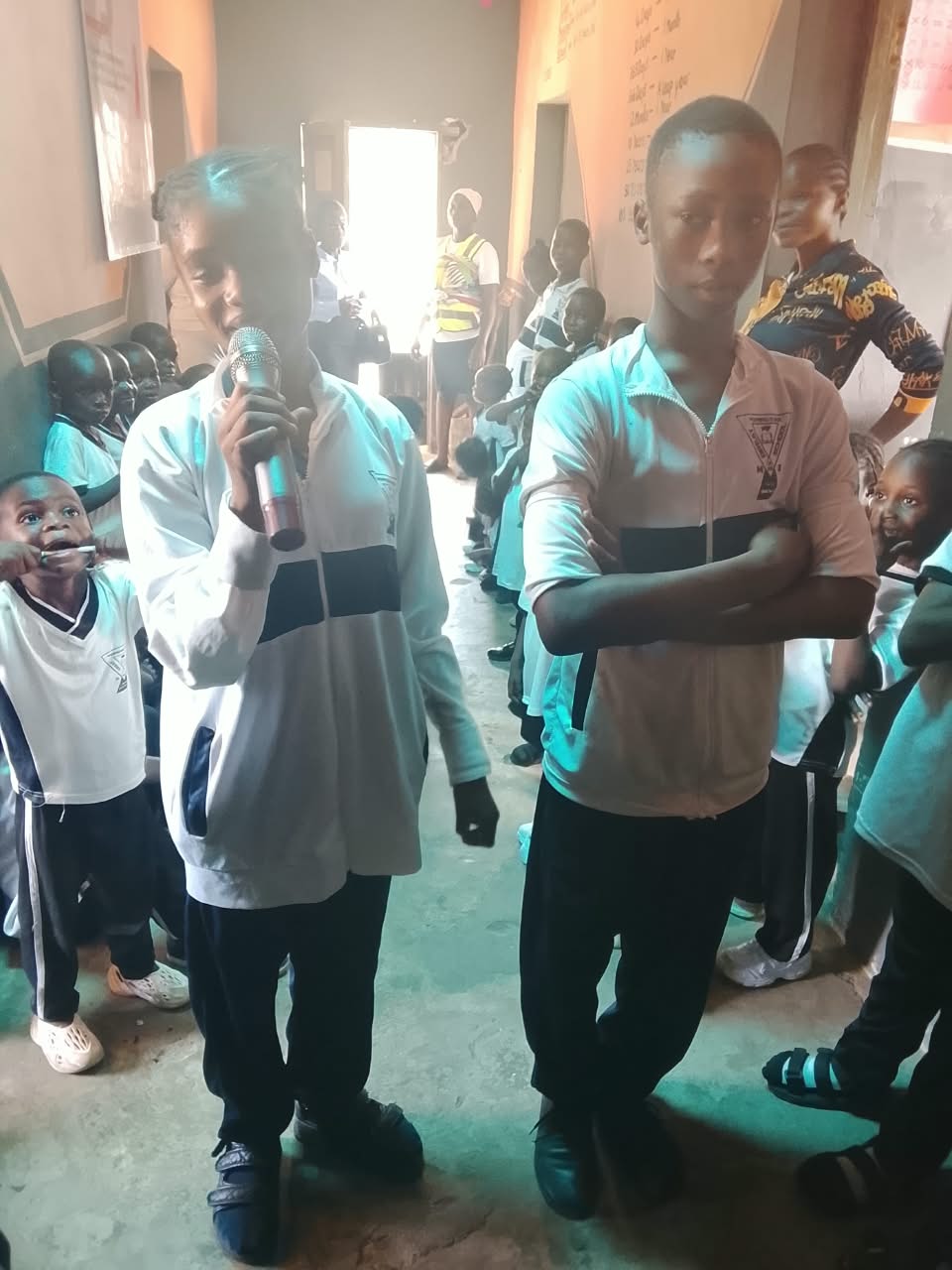 A student speaking into a microphone during the OLIC school seminar, a classmate beside her and younger pupils around them